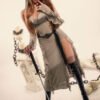 Cosplay model in a beige hooded dress with long brown hair, holding a sword and touching her lips, posed in a pale studio with statue-like decor in the background.