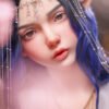 Close-up of a person with blue hair wearing a silver decorative chain headpiece and layered necklaces, with soft pink bokeh in the foreground.
