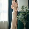 Woman in a beige evening gown leaning against a window, wearing strappy high heels indoors with green plants nearby.
