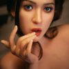 Close-up of a woman with blue eyes and glossy red lips, fingers near her mouth in a contemplative pose.