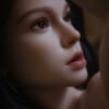 Close-up of a woman's face in profile, showing a brown eye with long lashes, arched eyebrow, smooth skin and pink lips in soft lighting.