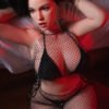 Woman in a black fishnet bodysuit posing on a bed with red lighting, showcasing a glamorous pose and jewelry (bracelet, hoop earring).