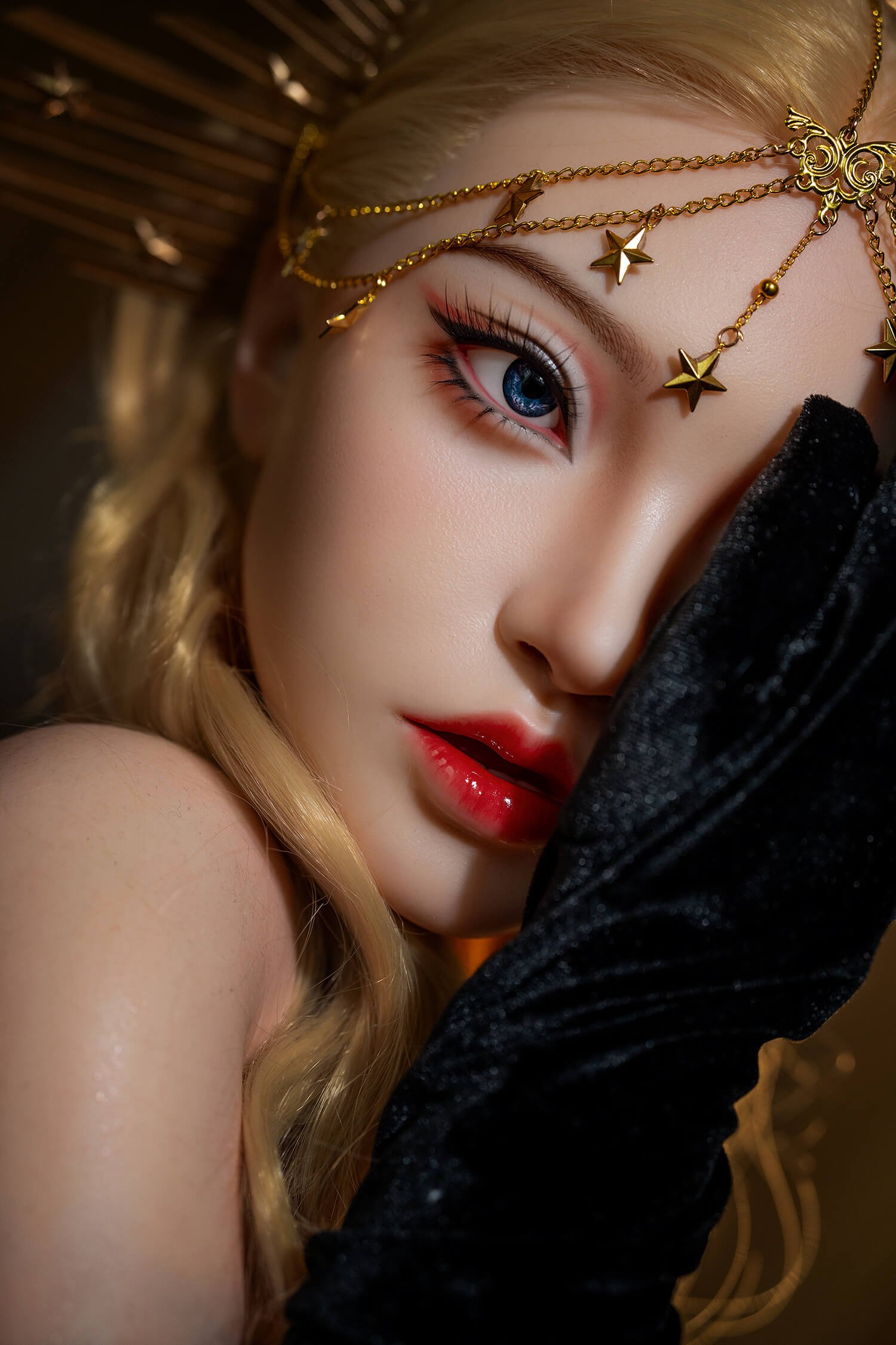 Close-up of a woman wearing a gold chain headpiece, blue eye makeup, and red lips, with a black velvet garment partially covering her face.