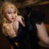 Model in a black feathered gown reclining, wearing a gold sunburst headpiece and bold red lips.