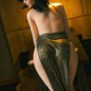 Woman with short black hair, back facing the camera, topless from the back and wearing snake-print pants and a gold chain, standing at a wooden counter in warm lighting.