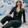 Woman in a black jumpsuit sits on snowy ground with icy mountains behind her; white fur hat and sunglasses hanging from her chest.